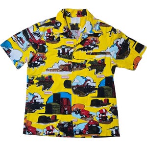 Puede incluir: Camisa amarilla de manga corta con un estampado repetido de maquinaria agrícola roja, azul y negra, incluyendo tractores y camiones. La palabra "Hessiton" está impresa en varias etiquetas rectangulares. Cuello camisero.