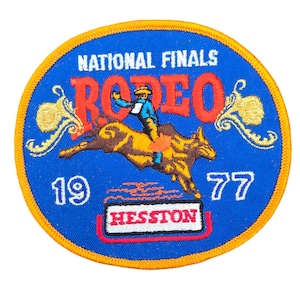 May include: A circular patch with a blue background and a yellow border. The patch features the words "NATIONAL FINALS RODEO" in white and red, with a cowboy riding a bull. The year "1977" and the word "HESSTON" are also displayed.