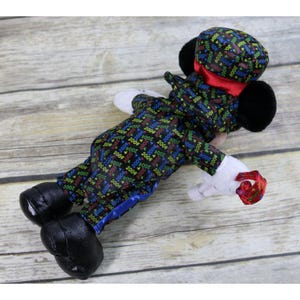 May include: A black plush Mickey Mouse dressed in a tuxedo with a blue satin trim and a black top hat. The tuxedo is covered in a repeating pattern of the year 2004 in various colors. Mickey Mouse is holding a red and gold party horn.