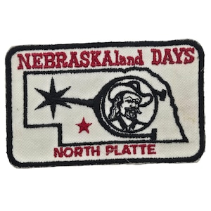 May include: A rectangular embroidered patch with a white background and black border. It features the red text NEBRASKALand DAYS and NORTH PLATTE. The design includes a black outline of Nebraska, a black star, and a black circular logo with a cowboy.