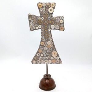 Könnte beinhalten: Ein graues Kreuz aus Knöpfen in verschiedenen Weiß-, Beige- und Brauntönen. Das Kreuz ist auf einem braunen Metallständer montiert.