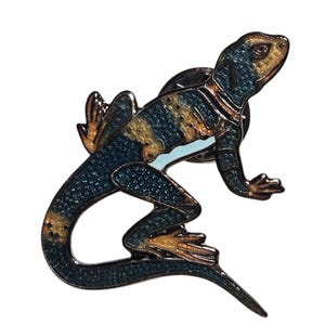 May include: A detailed enamel pin in the shape of a lizard. The lizard is primarily dark blue with gold and brown accents. The pin has a shiny, metallic finish and is set against a white background.