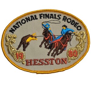 May include: An oval embroidered patch with a tan background and gold border. It features the text NATIONAL FINALS RODEO, 1980, and HESSTON. The design depicts two riders on brown horses roping a brown and yellow bull, all in colorful thread.
