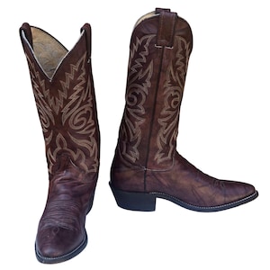 Puede incluir: Un par de botas vaqueras de cuero marrón con punta afilada, tacón de bloque y patrones de costura en color canela a lo largo de la caña alta. Las botas tienen un acabado desgastado y tiradores en la parte superior para facilitar su uso.