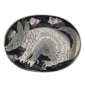 May include: An oval belt buckle featuring a silver-toned armadillo silhouette with etched scale details. The armadillo is set against a background of dark, iridescent mother-of-pearl inlay pieces that show flashes of purple, green, and pink.