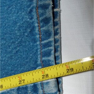 Puede incluir: Primer plano del borde inferior de un par de pantalones vaqueros azules con una cinta m&eacute;trica amarilla que muestra 27 pulgadas (68,58 cm).