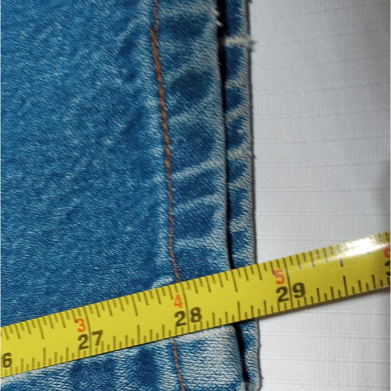 Puede incluir: Primer plano del borde inferior de un par de pantalones vaqueros azules con una cinta m&eacute;trica amarilla que muestra 27 pulgadas (68,58 cm).