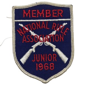May include: A vintage blue and white embroidered patch with the text "MEMBER NATIONAL RIFLE ASSOCIATION JUNIOR 1968". The patch features two crossed rifles and a white border.