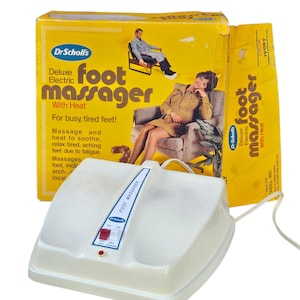 May include: Vintage Dr. Scholl's Deluxe Electric Foot Massager with Heat. The white foot massager has an on/off switch and a red indicator light. The box is yellow with illustrations of people relaxing and text that reads "For busy, tired feet!"
