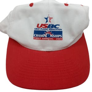 May include: A white and red baseball cap featuring the text "USBC Great Plains Association Championships First Annual 2005". The cap has a red peak and red embroidered dots on top.