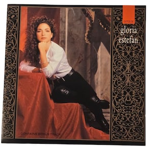 May include: Album cover featuring Gloria Estefan. She is seated, wearing a white shirt and black pants, with her hand under her chin. The album title "Exitos de Gloria Estefan" is visible, along with the text "Contains Bonus Track". The cover has a decorative border.