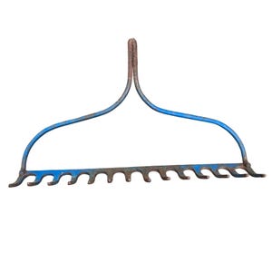 Puede incluir: Un rastrillo de jardín antiguo con un marco de metal pintado de azul. El rastrillo presenta un mango curvo y una hilera de púas. El metal muestra signos de edad y desgaste, con algo de óxido visible.