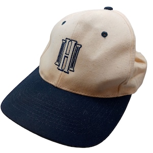 Puede incluir: Una gorra de béisbol beige con visera negra y una letra "H" bordada en azul marino en la parte delantera.