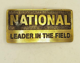 May include: A gold-coloured metal belt buckle with the word "NATIONAL" in large, raised letters and the phrase "LEADER IN THE FIELD" in smaller letters below.