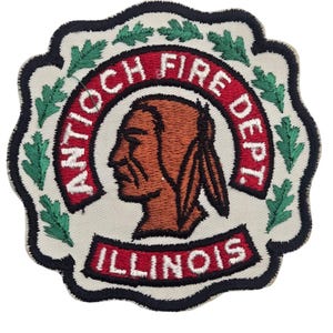 Op de afbeelding: Een ronde geborduurde patch met een geschulpte rand, voorzien van een bruin profiel van een persoon met veren in het haar. De tekst ANTIOCH FIRE DEPT. en ILLINOIS staat in wit op een rode achtergrond, omringd door groene eikenbladeren.