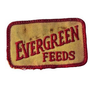 May include: A rectangular embroidered patch with a tan background and a dark red border. The text EVERGREEN FEEDS is stitched in dark red thread across the center. The patch shows signs of wear with small brown spots on the tan fabric surface.