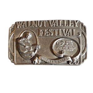 May include: A silver-coloured metal belt buckle with the words "Walnut Valley Festival" and "Winfield, Kansas" engraved on it. The buckle features a musician playing a guitar and the text "Home of the National Flat-Pick Guitar Championship."