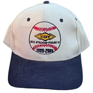 May include: A baseball cap with a beige crown and a navy blue brim. The cap features a baseball design with red stitching and the text "EBY ECK STADIUM - PHASE IV 1999-2000" in black and red.