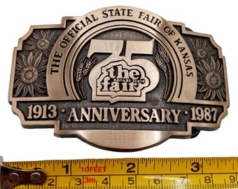 May include: A bronze belt buckle featuring the text "The Official State Fair of Kansas" and "1913 Anniversary 1987". The buckle depicts a large number "75" with the words "the fair" and "Kansas State" inside. The buckle is decorated with sunflowers.