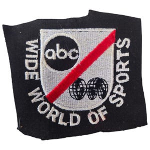 May include: Embroidered patch with the text "WIDE WORLD OF SPORTS" in white on a black background. The patch features a shield with the "abc" logo, two globe graphics, and a red diagonal stripe.