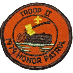 May include: Round orange patch with brown trim featuring the text "TROOP II HONOR PATROL 1979". The patch has a design of a log, axe, and fleur-de-lis. The patch is likely for a scouting organization.
