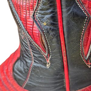 May include: Close-up of a cowboy boot with a red and black design. The boot features a red, textured vamp and a black shaft with decorative stitching. A red stripe runs down the back of the boot.