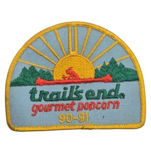 May include: A semi-circular embroidered patch featuring a yellow sun rising over green mountains and a red canoe. The text reads trail's end, gourmet popcorn, and 90-91 in yellow and red thread against a light blue background with a yellow border.