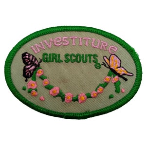 May include: Green oval embroidered patch with the words "Investiture Girl Scouts" in pink and green lettering. The patch features a pink butterfly and a yellow butterfly with pink accents, surrounded by a garland of pink flowers.