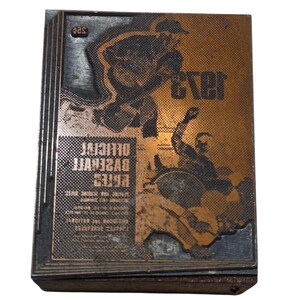 Puede incluir: Bloque de impresión vintage con temática de béisbol. El bloque presenta una superficie de color cobre con una ilustración de jugador de béisbol y la palabra "ever". También es visible el texto "OFFICIAL BASEBALL RULES".