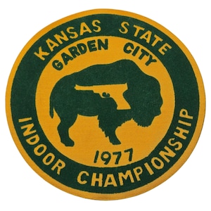 May include: A circular patch with a yellow background and green text and design. The text reads "KANSAS STATE GARDEN CITY INDOOR CHAMPIONSHIP 1977." A green silhouette of a bison with a gun superimposed on it is in the center.