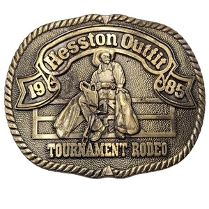Op de afbeelding: Een gouden gesp met een rodeo-thema. Het toont een cowboy die een wild paard berijdt, met de tekst "Heston Outfit 1968-1985" en "Tournament Rodeo" rond de rand.