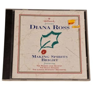 May include: A CD case featuring Diana Ross's "Making Spirits Bright" album, presented by Hallmark. The cover has a holly leaf design and text in blue and red. A yellow "SALE" sticker is on the upper right.