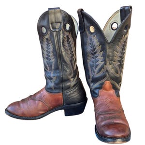 Könnte beinhalten: Ein Paar Cowboy-Stiefel mit zweifarbigem Design. Die Stiefel haben einen braunen Fuß und einen schwarzen Schaft mit dekorativen Nähten. Die Stiefel haben einen klassischen Western-Stil mit spitzer Zehenpartie und gestapeltem Absatz.