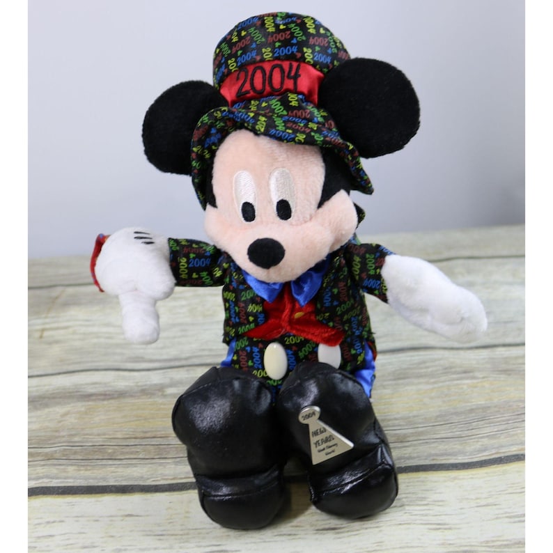 May include: A plush Mickey Mouse wearing a black and multi-colored tuxedo with a top hat that says "2004" on it. The plush is sitting down with its arms outstretched. The plush has a tag that says "Happy New Year's 2004".