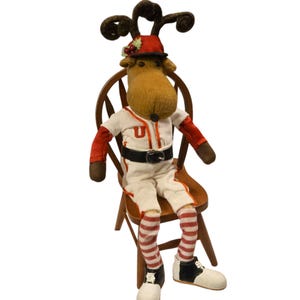 Könnte beinhalten: Ein Plüsch-Rentier, das eine rot-weiße Baseballuniform mit roter Mütze und schwarzem Gürtel trägt. Das Rentier sitzt auf einem Holzstuhl mit rot-weiß gestreiften Strümpfen und schwarz-weißen Schuhen.