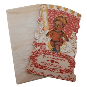 Peut inclure: Carte de Saint-Valentin vintage représentant un garçon chérubin aux cheveux roux et vêtu d'une tenue rouge et blanche, tenant une colombe. La carte est de couleur rouge, blanche et or et présente un motif floral. Le texte sur la carte est "To My Valentine, Valentine with charms so sweet, I lay this offering at your feet."