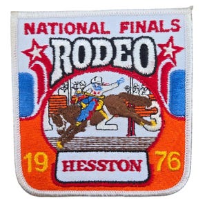 May include: Embroidered patch with the words "NATIONAL FINALS RODEO" in white lettering. The patch features a rodeo scene with a cowboy riding a bucking bronco. The text "HESSTON 76" is also included, with a red, white, blue, and orange color scheme.