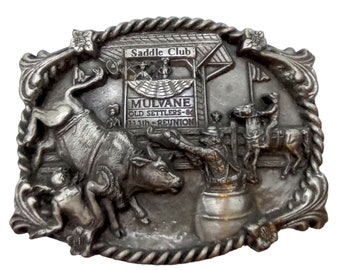 Puede incluir: Una hebilla de cinturón plateada con un diseño 3D que representa una escena de rodeo. La hebilla presenta un toro que lanza a un vaquero, un vaquero montando un caballo y un letrero que dice "Mulvane Old Settlers - 84 113th Reunion Saddle Club".