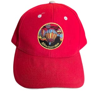 May include: Red baseball cap with an embroidered circle patch featuring hot air balloons and the text "Albuquerque".