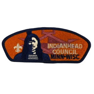 Puede incluir: Un parche bordado de color naranja y azul con el texto "Indianhead Council Minn-Wisc". El parche presenta una cabeza de nativo americano con un tocado de plumas y un símbolo de scout.