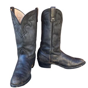 May include: A pair of worn black leather cowboy boots. The boots have a pointed toe, decorative stitching, and a tall shaft. The leather shows signs of wear and tear, with scuffs and creases. The boots are set against a white background.