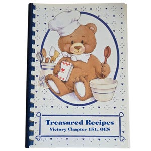 Op de afbeelding: Een spiraalgebonden kookboek met een illustratie van een teddybeer als chef-kok op de omslag. De beer draagt een koksmuts en houdt een lepel en een receptenboek vast. De titel "Treasured Recipes" staat eronder, met een blauw en wit hartjespatroon.