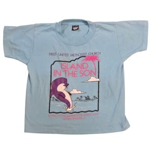 May include: A light blue short-sleeved t-shirt featuring a graphic of a purple dolphin jumping out of the water. The text reads FIRST UNITED METHODIST CHURCH, ISLAND IN THE SON, LIVING IN THE WARMTH OF GOD'S LOVE, and 1 CORINTHIANS 13 in pink and black.