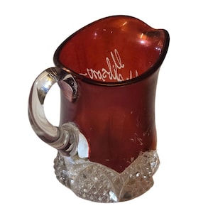 May include: A ruby red glass pitcher with a clear handle and a diamond-cut base. The interior of the pitcher has white cursive text. The pitcher is likely vintage and suitable for serving beverages.