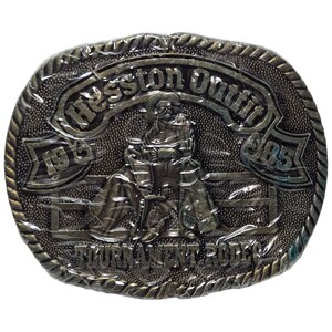 May include: Oval-shaped, antique-style belt buckle with a detailed relief of a rodeo scene. The buckle features the text "Hesston Outfit" and "Tournament Rodeo" with the years "1976" and "85" incorporated into the design. The buckle has a textured background and a rope-like border.
