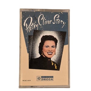 Op de afbeelding: Een vintage cassettebandje met "The Patsy Cline Story". De hoes toont een portret van Patsy Cline tegen een blauw-witte achtergrond. Het Decca Records-logo is zichtbaar. De cassette zit in een doorzichtige plastic hoes.