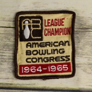 May include: A brown and beige embroidered patch with the text "League Champion" and "American Bowling Congress" and the years "1964-1965". The patch features a white bowling pin with the letters "ABC" in gold.