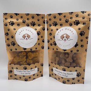 May include: Two brown paper bags with paw print designs. Each bag contains dog biscuits and has a label with the text "UPPY PUPPY PET BAKERY". The left bag contains pumpkin oat dog biscuits and the right bag contains peanut butter dog biscuits. The bags are labeled with the weight of the biscuits in ounces and grams.