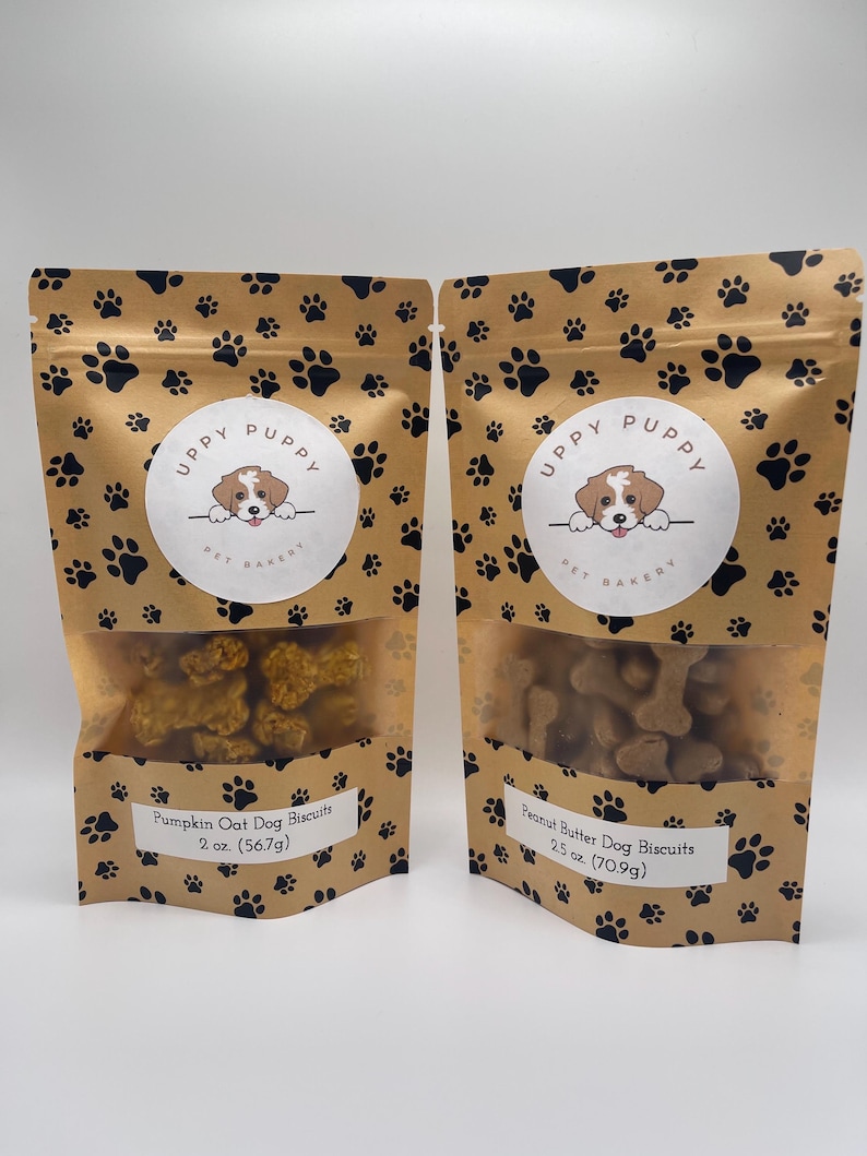 May include: Two brown paper bags with paw print designs. Each bag contains dog biscuits and has a label with the text "UPPY PUPPY PET BAKERY". The left bag contains pumpkin oat dog biscuits and the right bag contains peanut butter dog biscuits. The bags are labeled with the weight of the biscuits in ounces and grams.