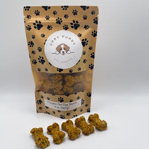 May include: A brown bag with a paw print pattern and the text "Uppy Puppy Pet Bakery" on the front. The bag contains pumpkin oat dog biscuits. There are six orange bone-shaped dog biscuits on a white surface.